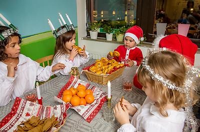 Five children sit at a table in Saint Lucy and Santa Claus costumes, eating lussekatter, gingerbread cookies, and oranges. 