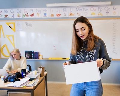 En elev håller ett föredrag inför klassen medans läraren sitter vid katedern bakom henne.