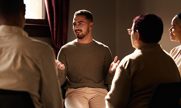 Dark-haired man in early middle age, dressed in light-coloured clothing, sits and leads a group discussion. He gestures as he speaks and is met by attentive gazes and smiles.