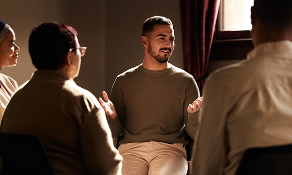 Dark-haired man in early middle age, dressed in light-coloured clothing, sits and leads a group discussion. He gestures as he speaks and is met by attentive gazes and smiles.