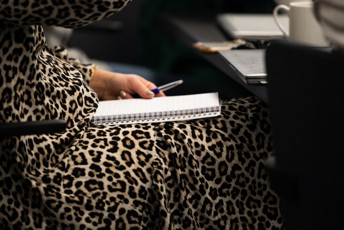 Close-up of a person holding a pen over a notebook in their lap. A laptop and a coffee mug can be glimpsed in the image.